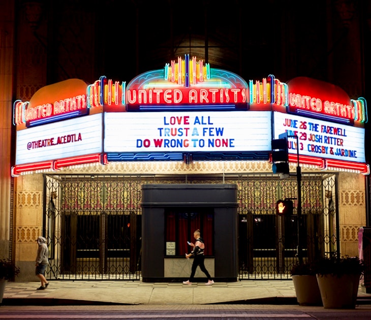 People walk in front of the Ace Hotel at night, which has an old-fashioned movie house marquee lit up in neon; Los Angeles neon
