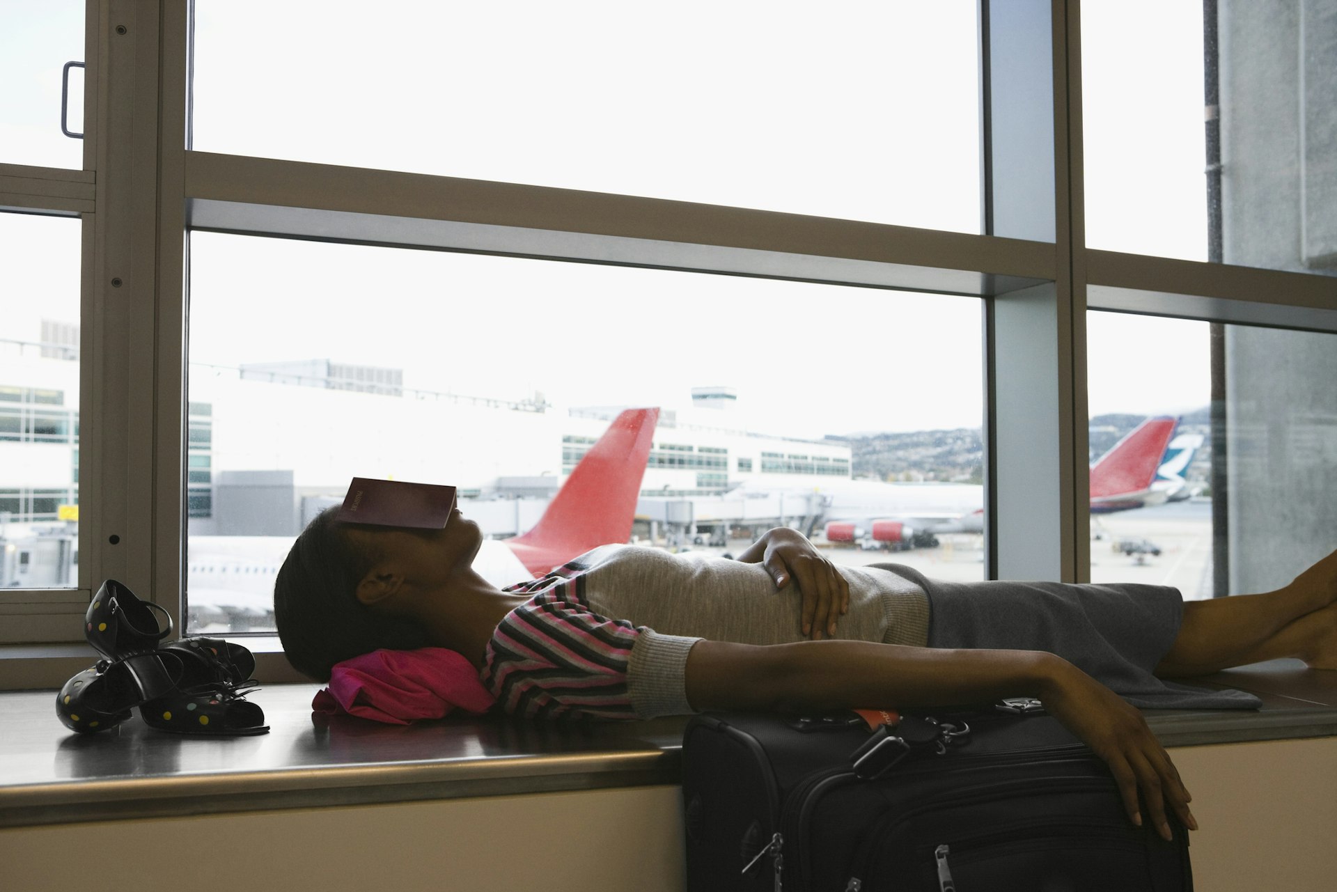 A woman with jet lag lies on her back sleeps on a large window sill. There is a open book laid across her face to block the light. One hand is folded over her stomach while another is outstretched over her suitcase. Her shoes are set just above her head on the sill. Outside the window are planes parked at gates.