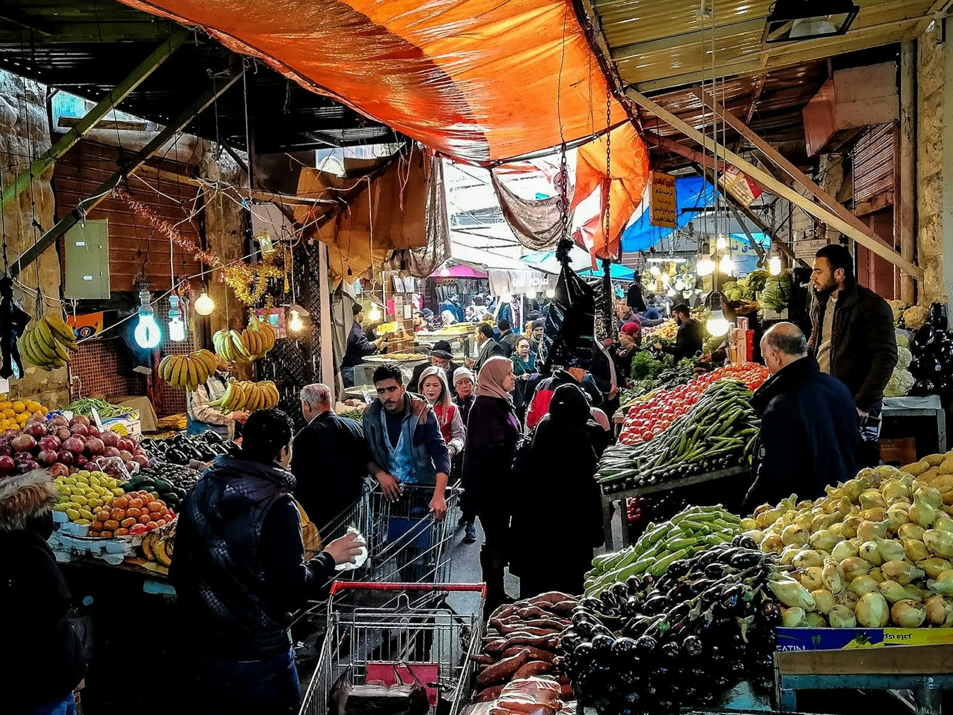 Fruit and vegetables are piled high on both sides of a narrow walkway under a covered market; shoppers push carts or carry bags, while sellers stand behind their goods on raised platforms; Free things to do in Amman, Jordan