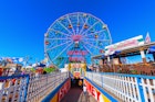 A colourful ferris wheel stands in front of a brilliant blue sky, with colourful railings and placards flanking the tunnel down to the wheel's entrace