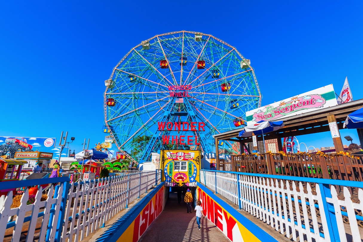 A colourful ferris wheel stands in front of a brilliant blue sky, with colourful railings and placards flanking the tunnel down to the wheel's entrace