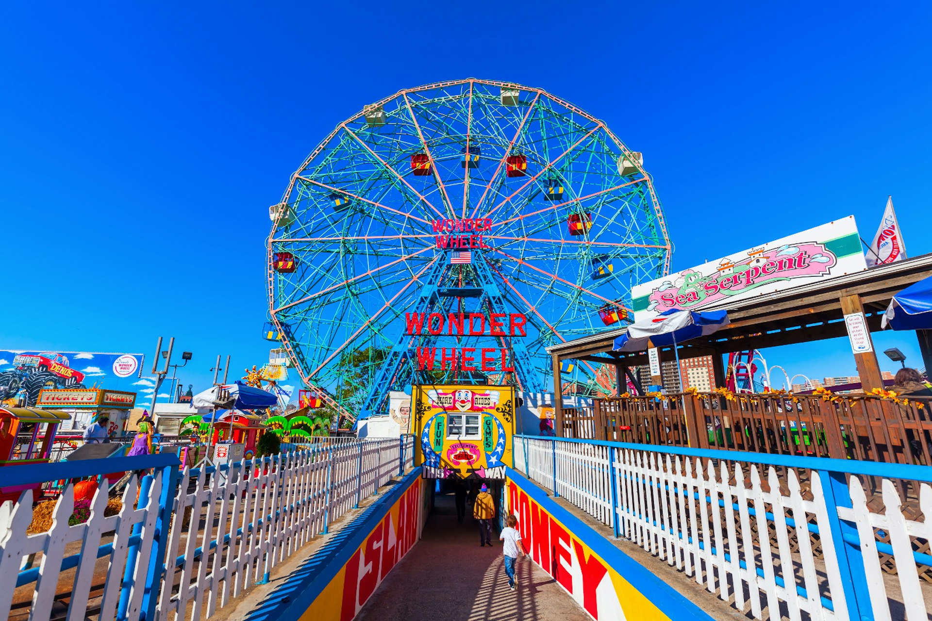 A colourful ferris wheel on Coney Island stands in front of a brilliant blue sky, with colourful railings and placards flanking the tunnel down to the wheel's entrace 