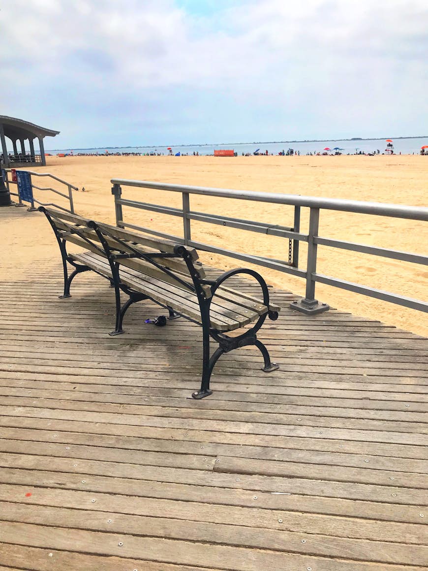 A wooden-and-iron bench sits on the boardwalk, with the empty sands of Brighton Beach in the background.