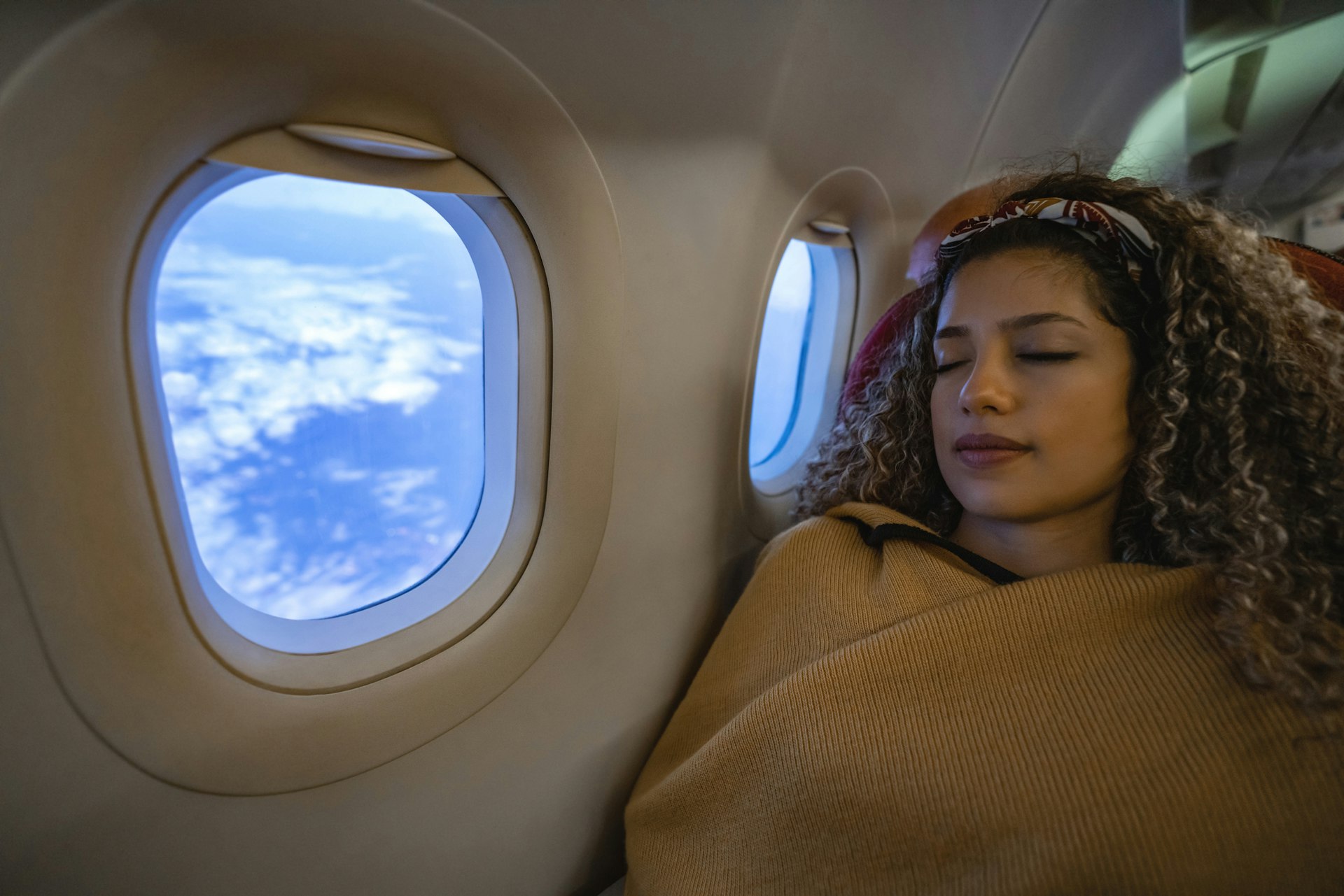 A woman sleeps under a blanket next to a plane window. The blind is up, revealing clouds below. Taking a longer first leg may help get extra sleep, which can reduce jet lag.