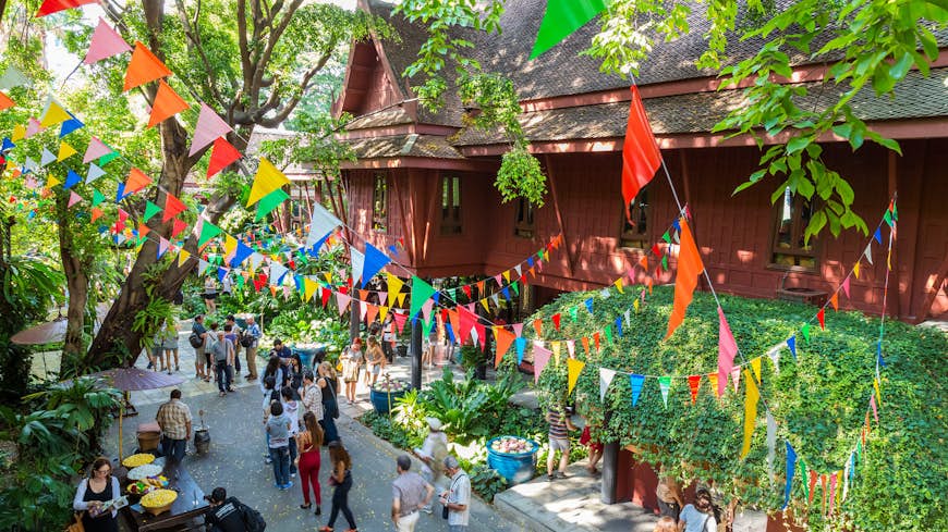 A stilted, maroon-coloured house, with a steep-tiled roof, sits among trees and numerous strings of colourful bunting; visitors crowd the walking paths around the home