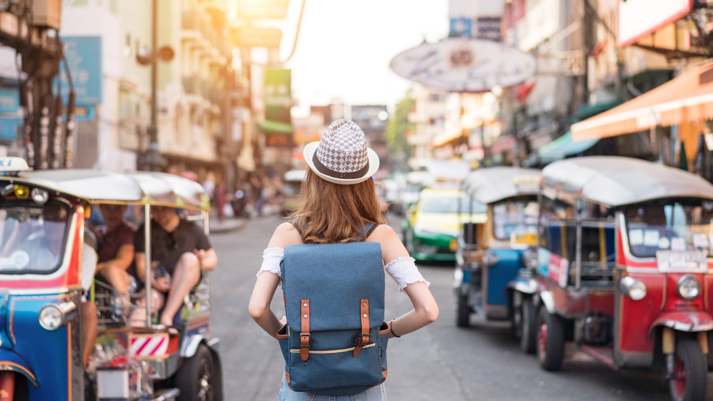 The back of a young woman walking along the pedestrian street in the evening in Bangkok, Thailand; travelers, tourists and tuktuks line the street.