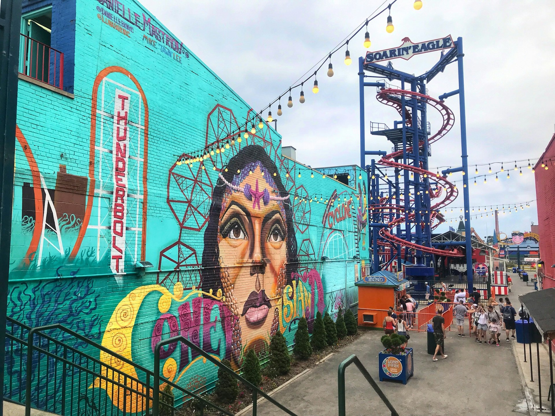 A vibrant street art mural on a woman's face covers he side of a brick wall that is coloured aquamarine; in the background is an amusement ride