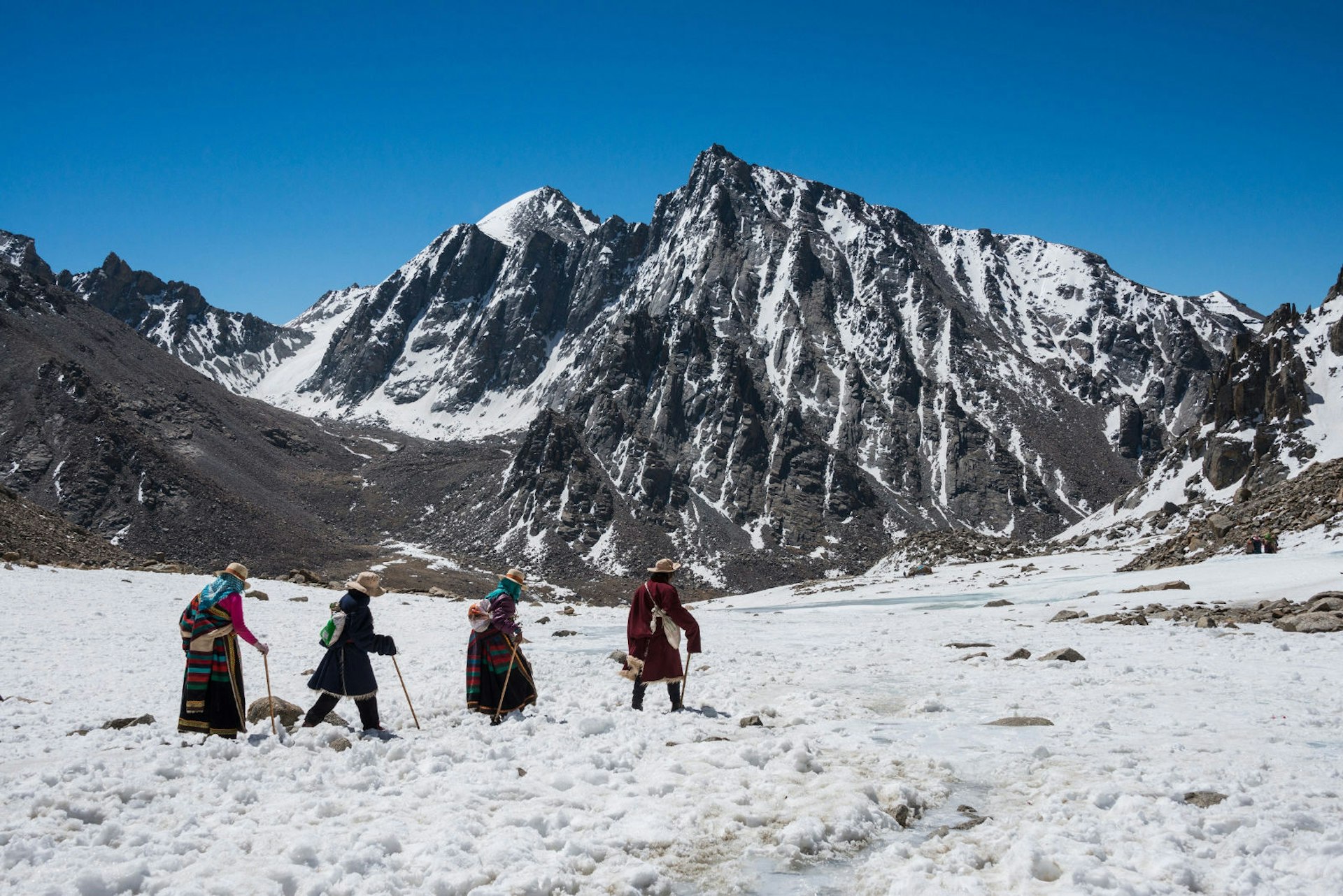 Four people dressed in heavy winter clothing walk through the snow with a snow-capped peak in the background and a clear blue sky.