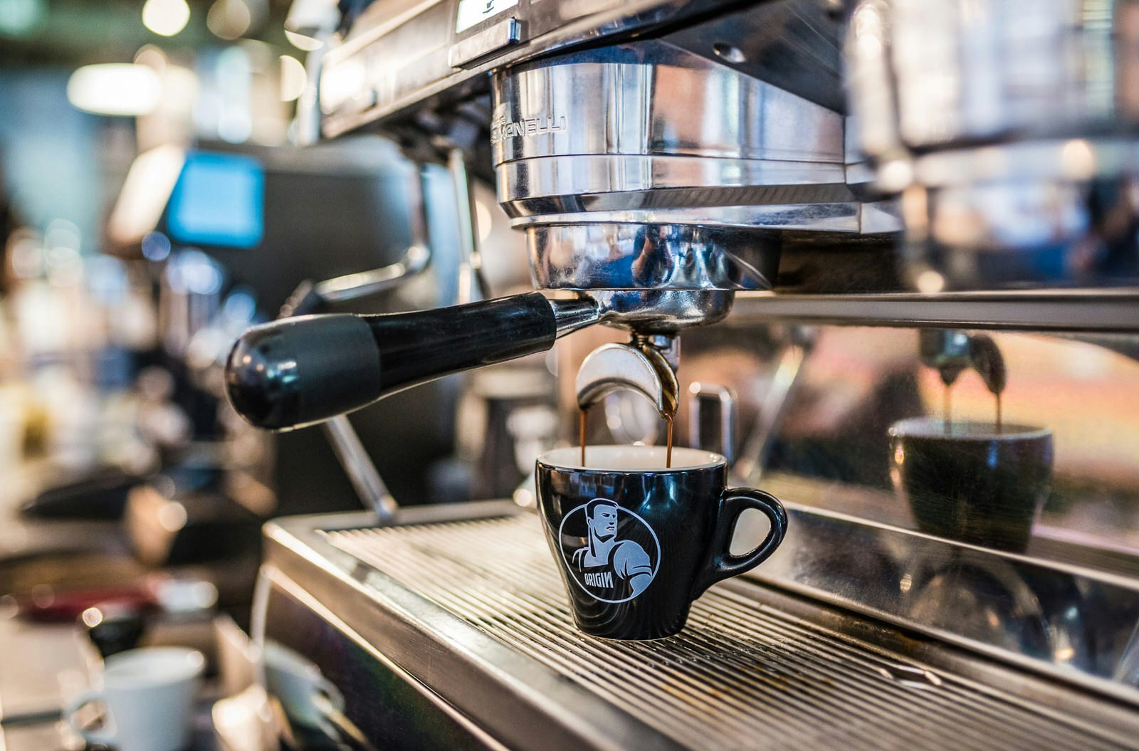 Espresso pours out of a large chrome coffee machine in two streams; both ball into a small black cup with an Origin logo (a barista)