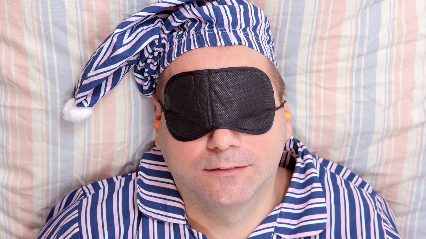 A middle-aged man dressed in blue and white striped pyjamas and matching hat reclines on a striped bedsheet with a black eye mask over his face and ear plugs.