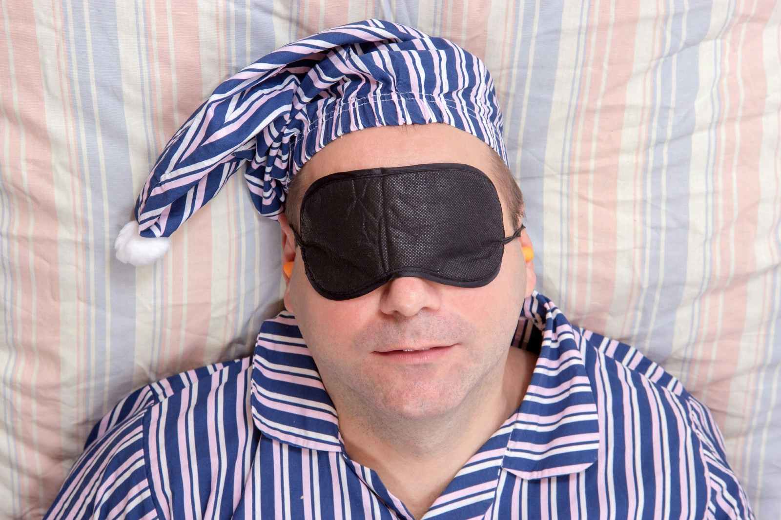 A middle-aged man dressed in blue and white striped pyjamas and matching hat reclines on a striped bedsheet with a black eye mask over his face and ear plugs.