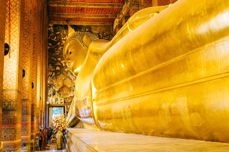 Within a small door at the distant end of a Thai temple (Wat Pho) stand a few visitors who are dwarfed by the gargantuan 46m-long Reclining Buddha statue; its feet are off the end of the picture, with its legs visible moving up towards its waste and towering head; the walls and ceiling of the temple are rich in gold, red hues and elegant designs.