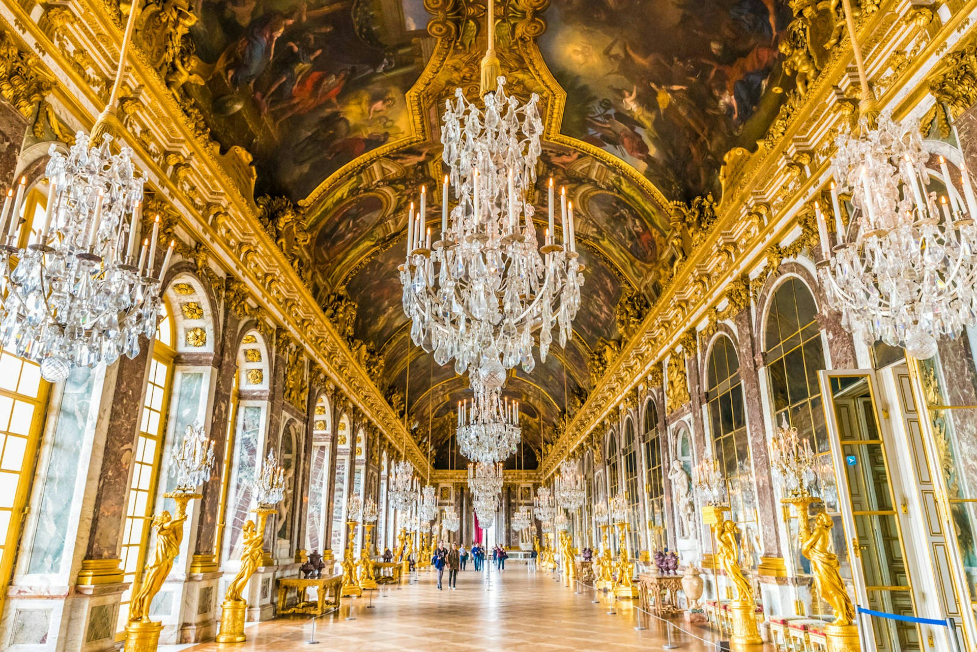 Interior fo the Château de Versailles: an extremely grand corridor with gold statues, huge windows and ornate, crystal chandeliers dangling from the ceiling