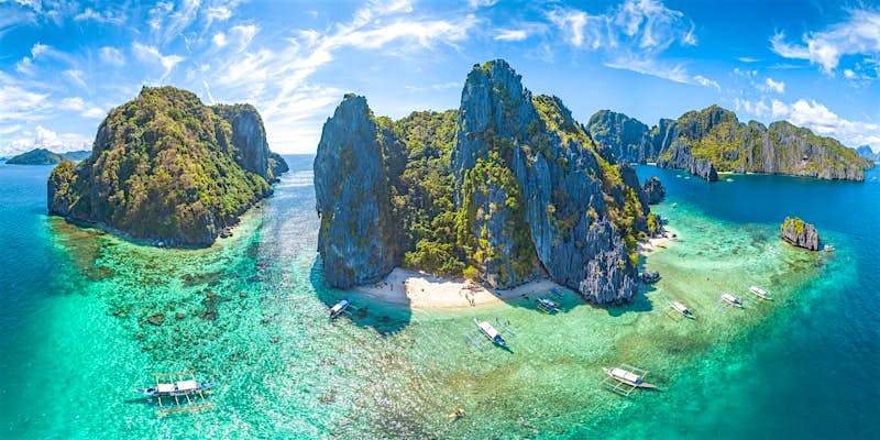 An aerial shot of El Nido, Palawan Province, Philippines. The small islands have very high rocky cliffs and lots of dense greenery. There are somewhite sandy beaches and a few boats moored near the islands. 