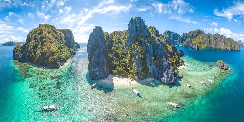 An aerial shot of El Nido, Palawan Province, Philippines. The small islands have very high rocky cliffs and lots of dense greenery. There are somewhite sandy beaches and a few boats moored near the islands.