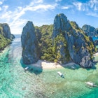 An aerial shot of El Nido, Palawan Province, Philippines. The small islands have very high rocky cliffs and lots of dense greenery. There are somewhite sandy beaches and a few boats moored near the islands.