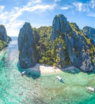 An aerial shot of El Nido, Palawan Province, Philippines. The small islands have very high rocky cliffs and lots of dense greenery. There are somewhite sandy beaches and a few boats moored near the islands.