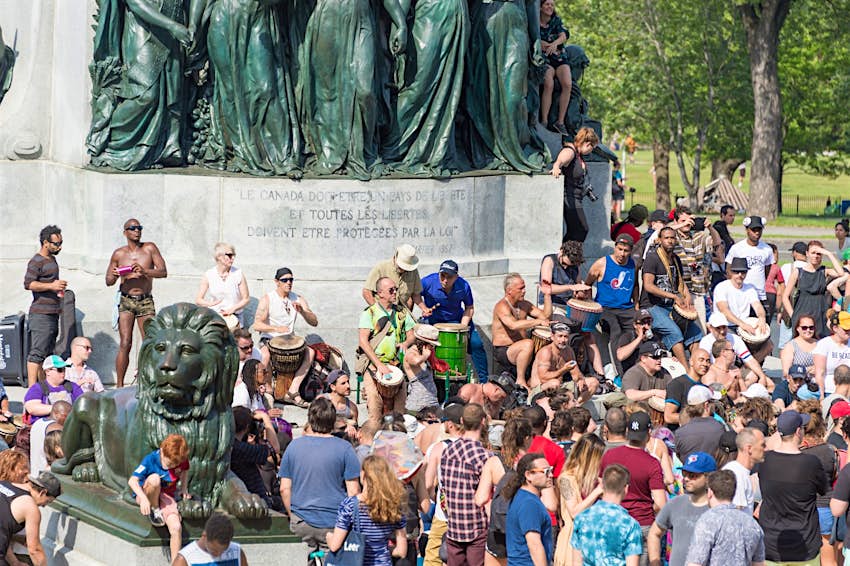 A large group of people play TamTams and other drums as other people dance and revel in the nice weather around a statue; free things to do in Montréal