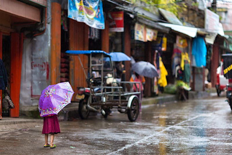 A little girl sheltering under a purple umbrella standing alone in the street in the rain in Banuae village, Philippines looking towards people sheltering in colorful shops