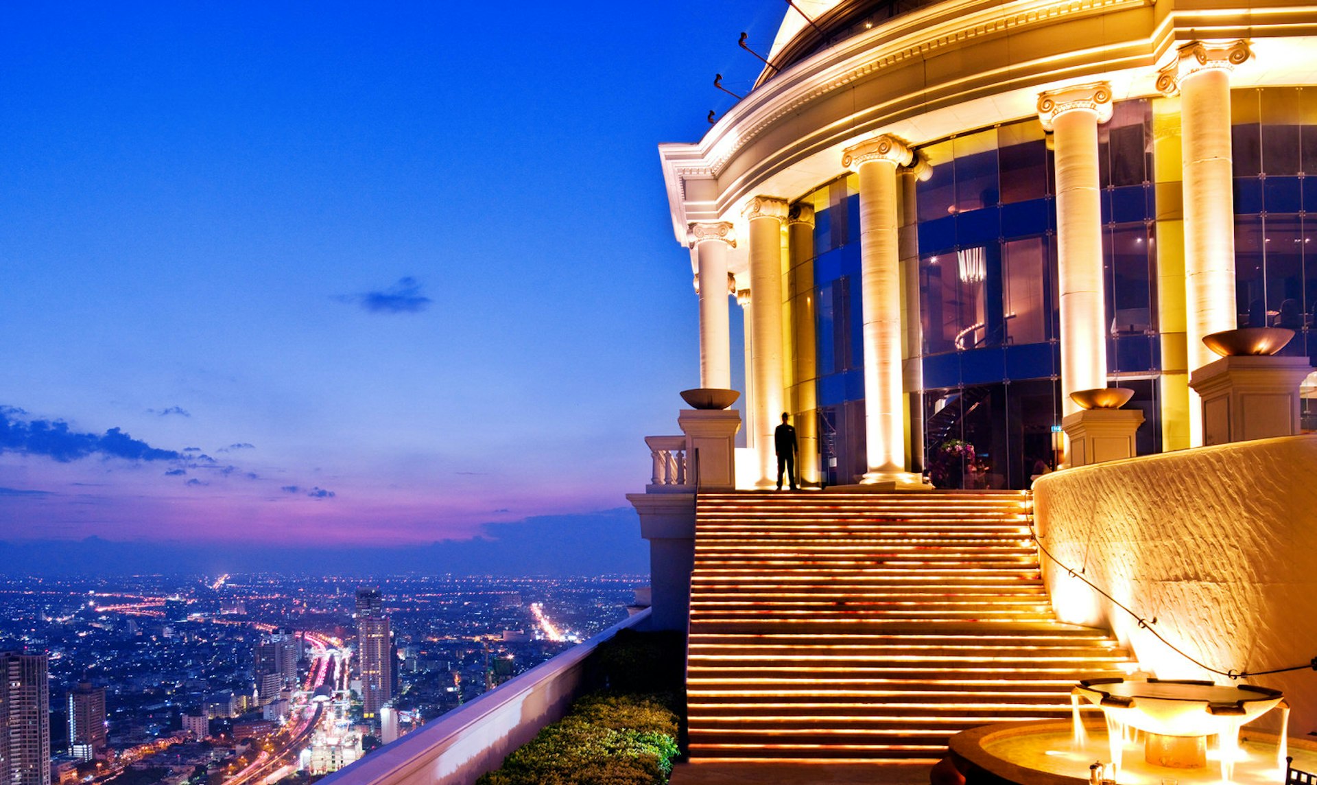 A Roman-looking circular stone building with large columns is lit up brilliantly from below; to the left, down in the distance is the teeming city of Bangkok