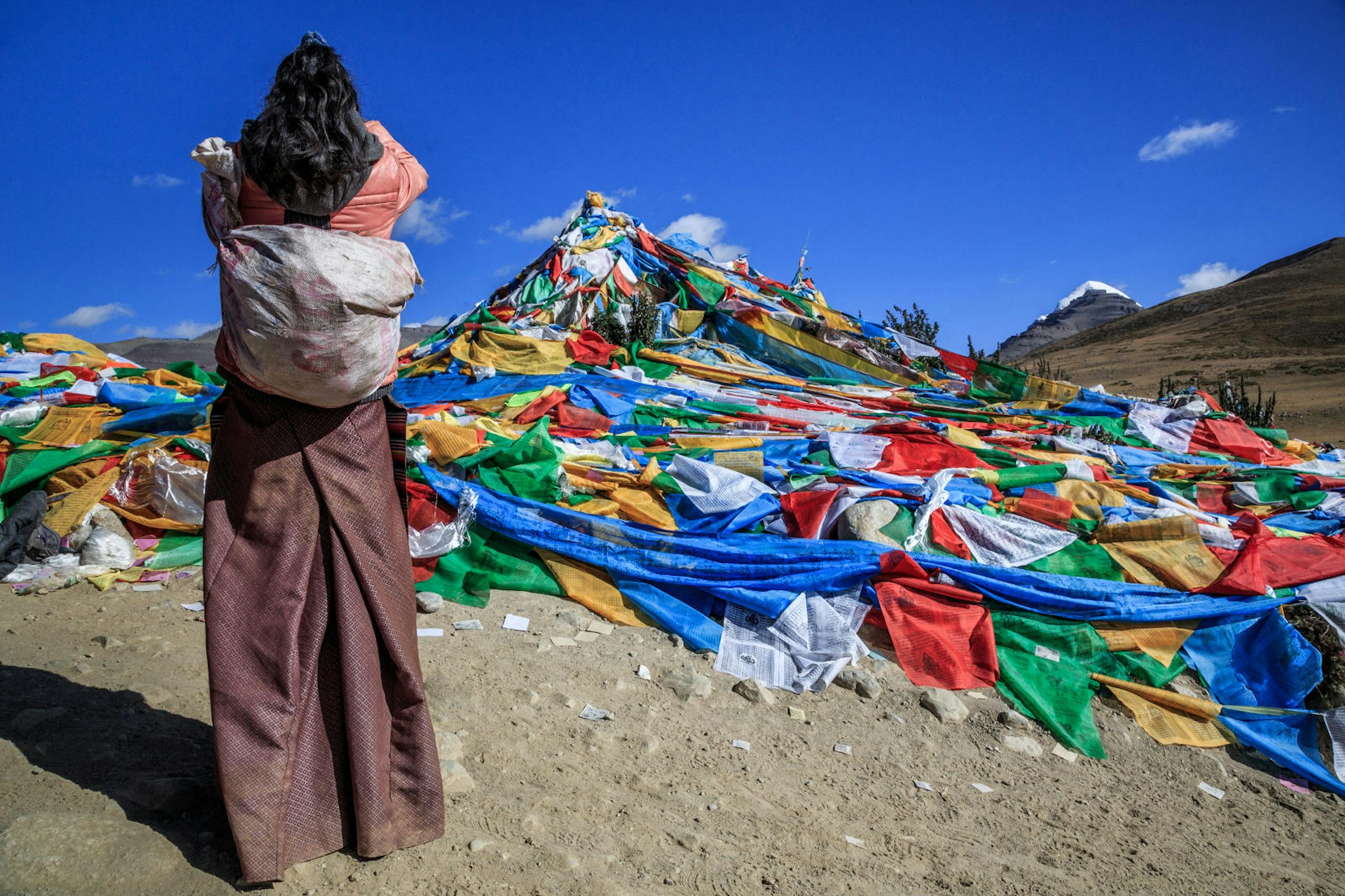 Multi-coloured Tibetan prayer flags cover a small hill. A woman is standing with her back to the camera praying; she has dark hair and is wearing a traditional long skirt, warm jacket and has a bag slung over her shoulder. The snow-capped peak of Mount Kailash is visible in the background.