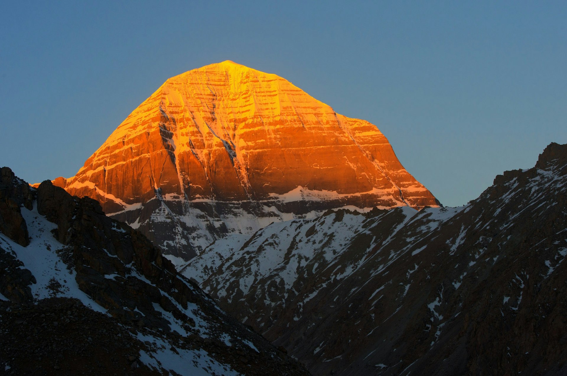 The peak of the mount Kailash seen in an orange light at dawn. The lower part of the mountain is in darkness and covered in snow.