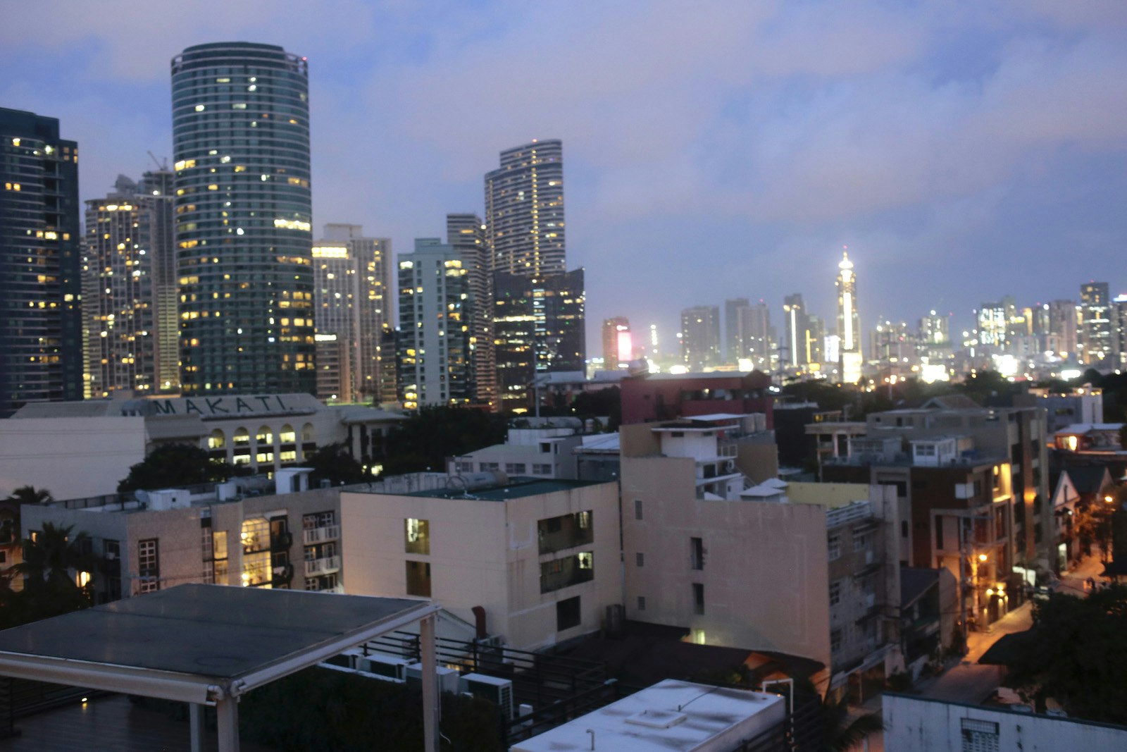 Rooftop view of Makait City at dusk with lit high-rises in the background and white cement buildings in the foreground