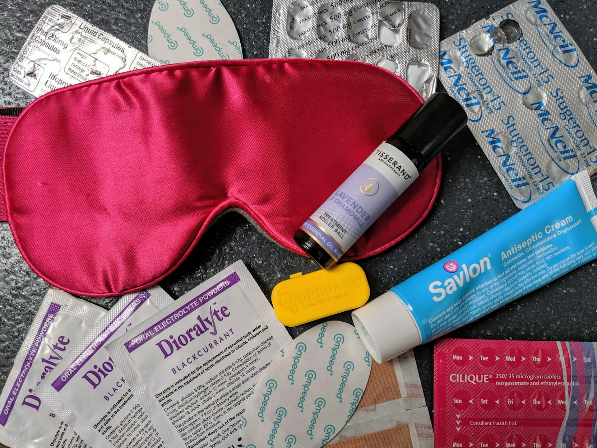 An arrangement of health essentials, including a pink sleep mask, antiseptic cream, painkillers, plasters and ear plugs