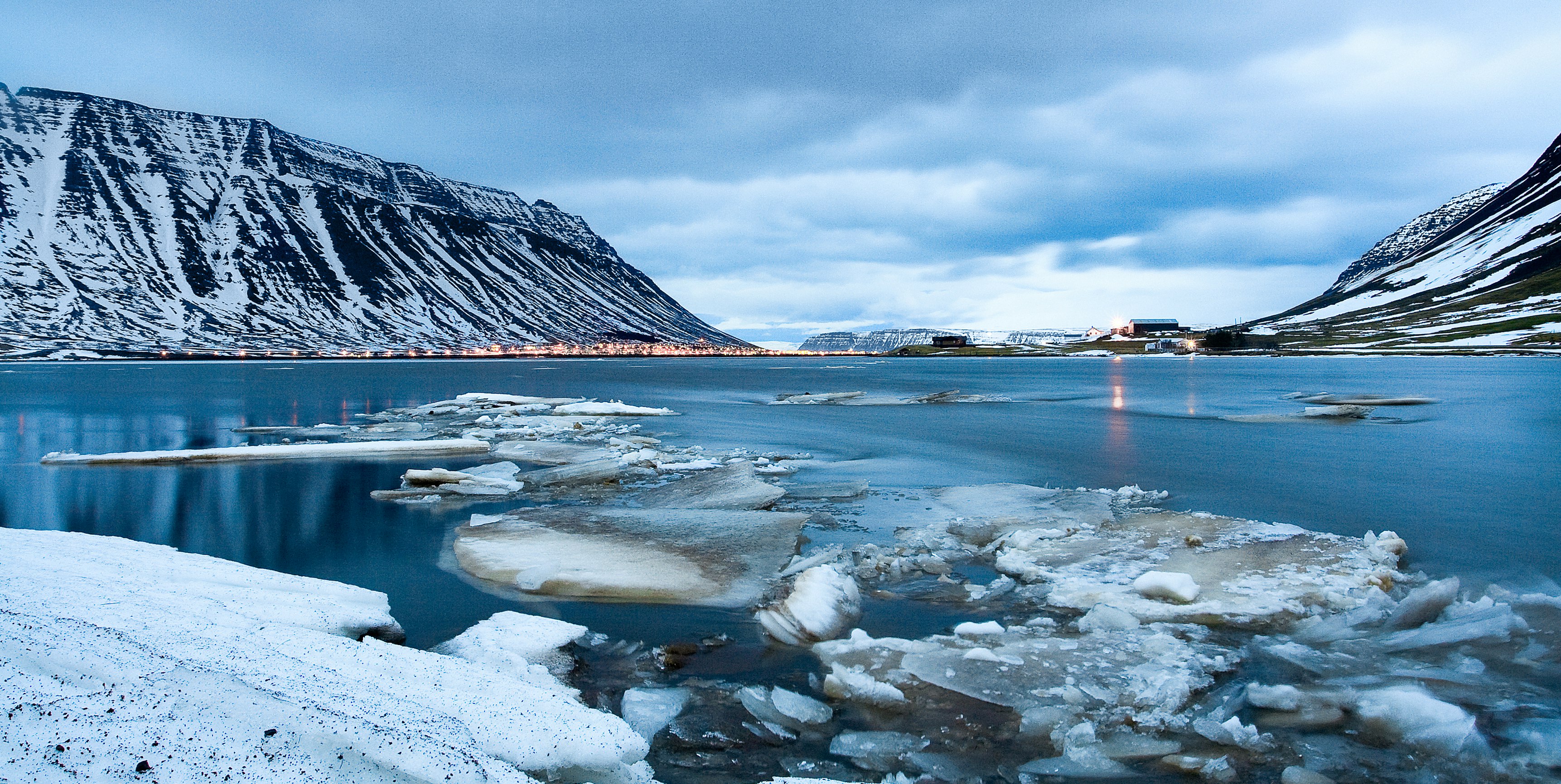 Winter afternoon in Ísafjörður, Westfjords; the fjord contains ice and the lights of the town are visible on the far side of the fjord.