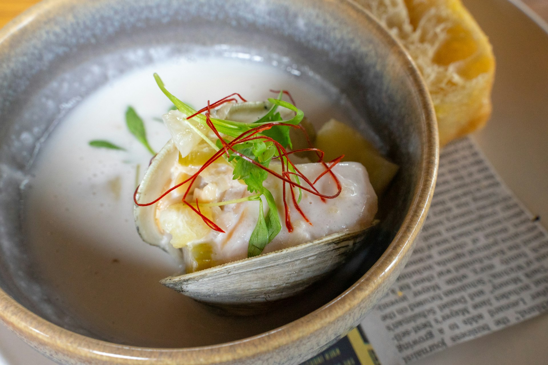 A glazed stoneware bowl of clam chowder at Union in Portland, Maine, features a light white broth against which micro greens and bright red saffron strands stand out as they drape of a clam still in-shell