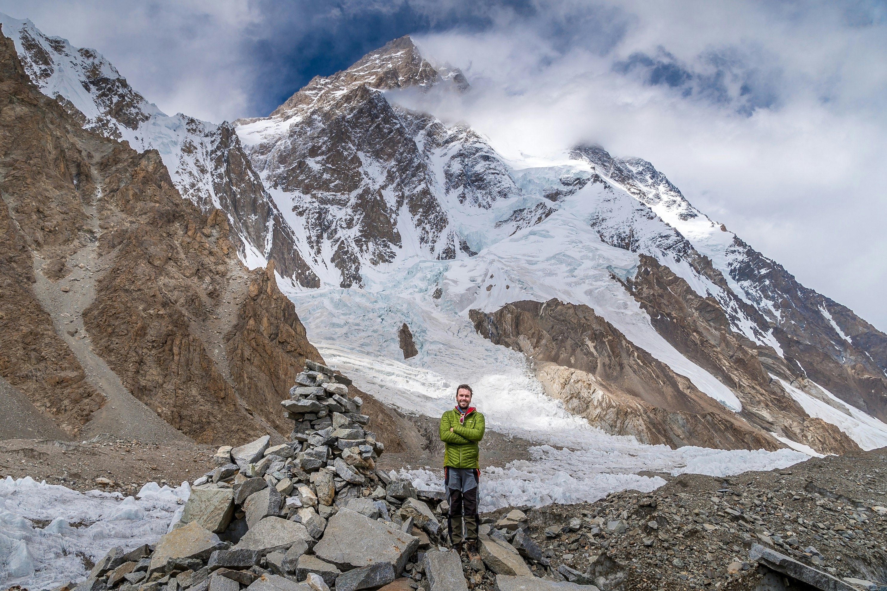 Peter, wearing a bright green trekking jacket, stands next to a small rock pile that marks K2 base camp. K2 is visible behind him, along with other, smaller, mountain peaks.