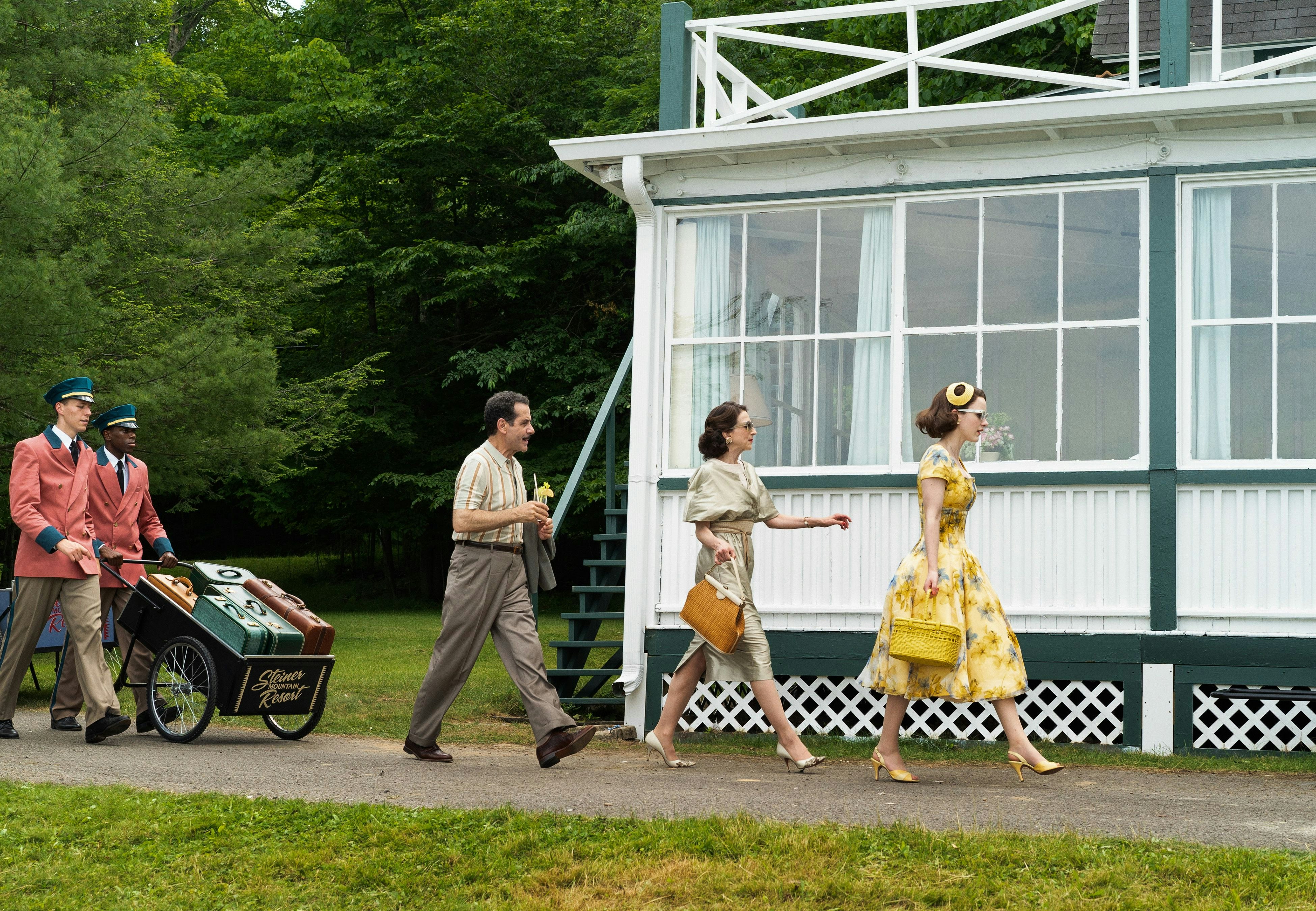 A still from season 2 of The Marvelous Mrs. Maisel, which shows Midge, her mother, and father walking single file in front of a white and forest green bungalow at a fictional Borscht Belt resort. Midge is wearing a yellow floral dress with matching shoes, handbag, and hat. Her mother is in a dove grey silk with matching shoes and a brown handbag. Her father is in dark khaki slacks and a neutral buttondown. They are followed by two porters in pink uniform coats and black hats pushing luggage in a black cart