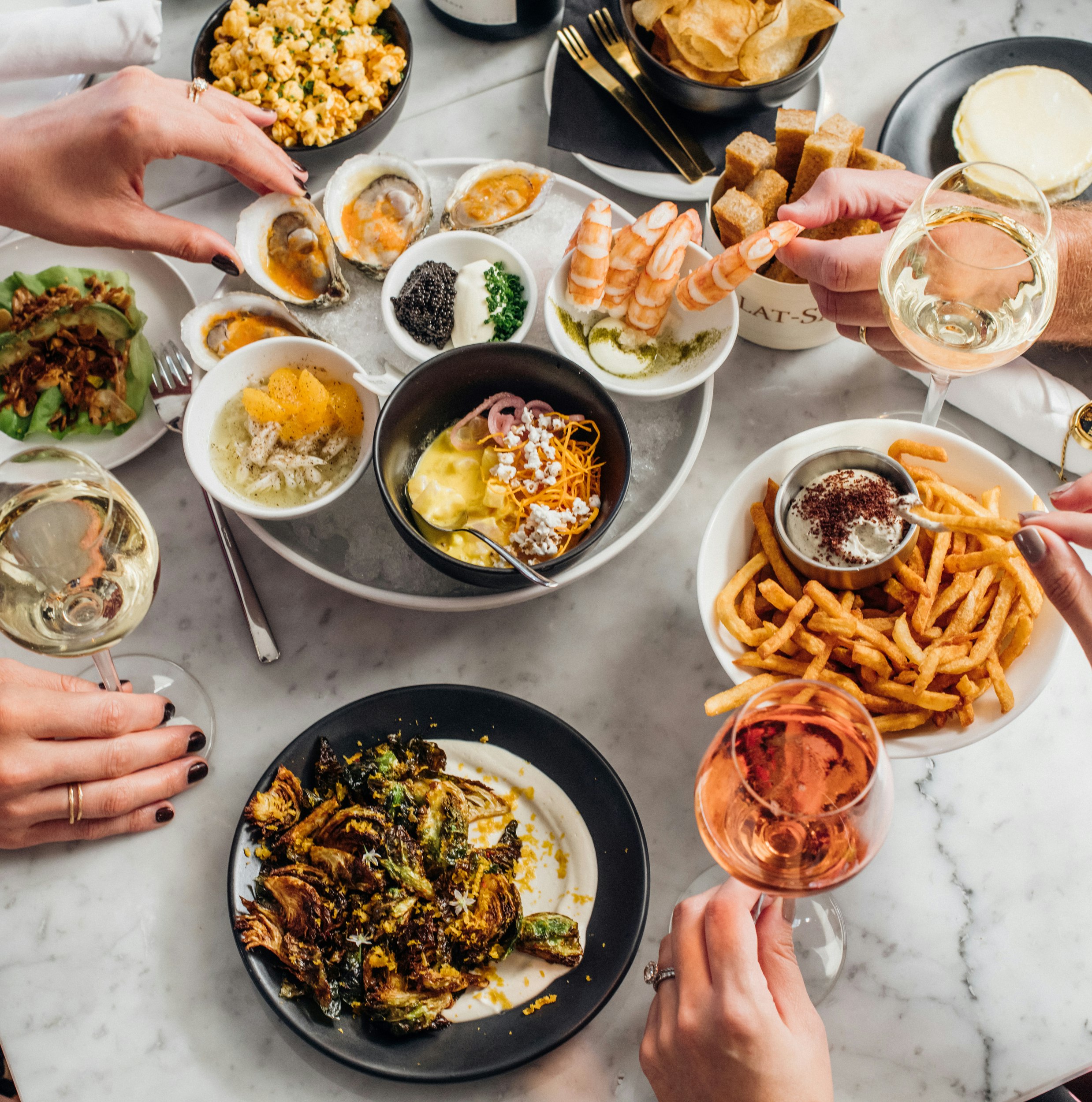A marble table is laden with plates of colorful food, from oysters on the half-shell to golden frites. At least two pairs of white, female hands reach for bites of food while holding full glasses of white and rose sparkling wines.