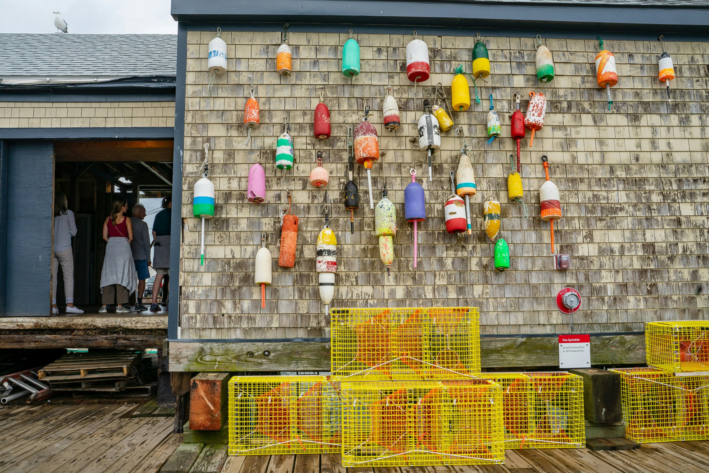 The worn wood shingles on the side of Luke's Lobster on the Portland pier is covered in colorful buoys over the wharf itself, where yellow lobster traps are stacked. To the left of the frame, up steps made from shipping palettes, stands a tour group with their backs to the camera 