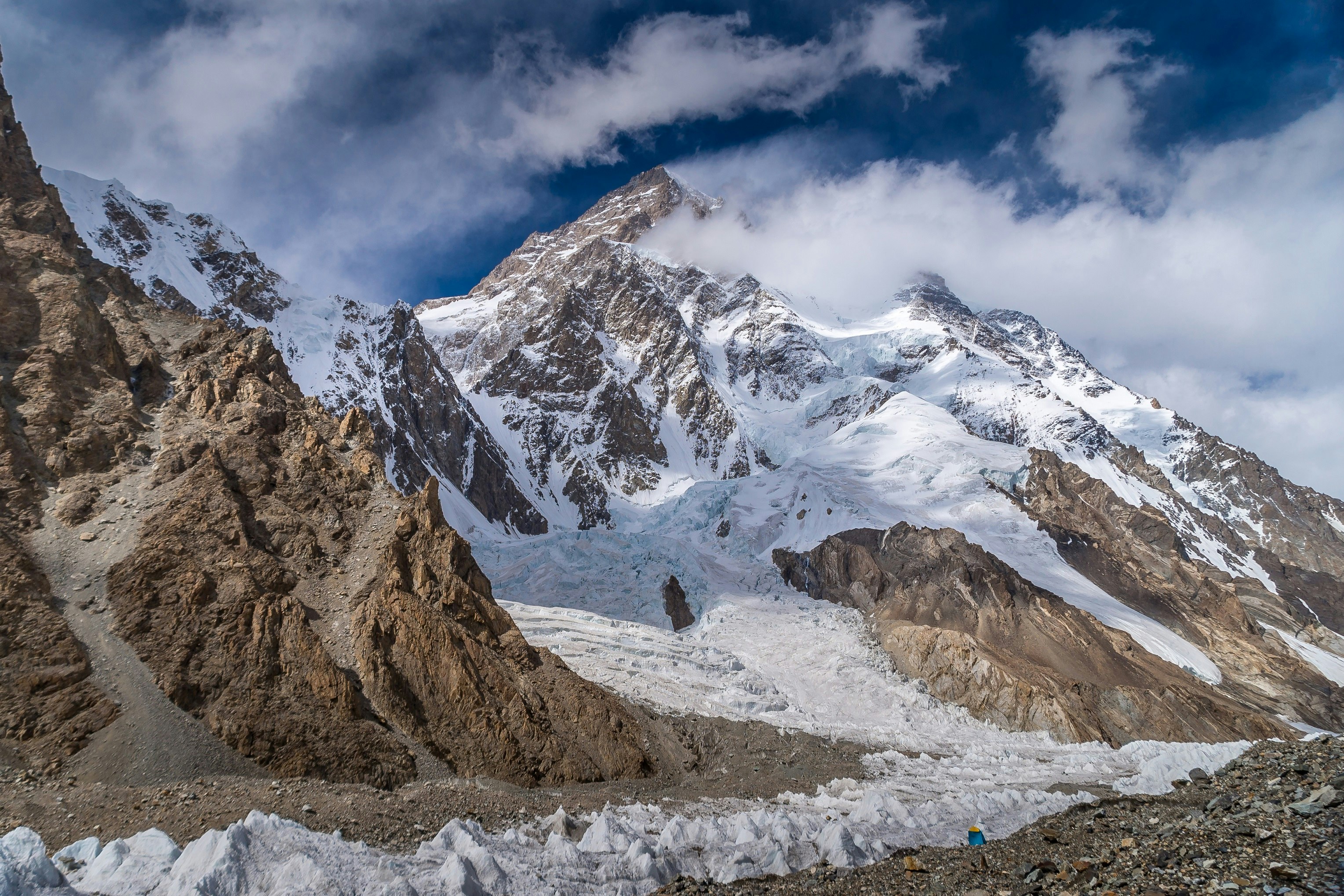 A view of K2 mountain from base camp. The rocky mountain is covered in snow and its peak is obscured slightly by cloud.