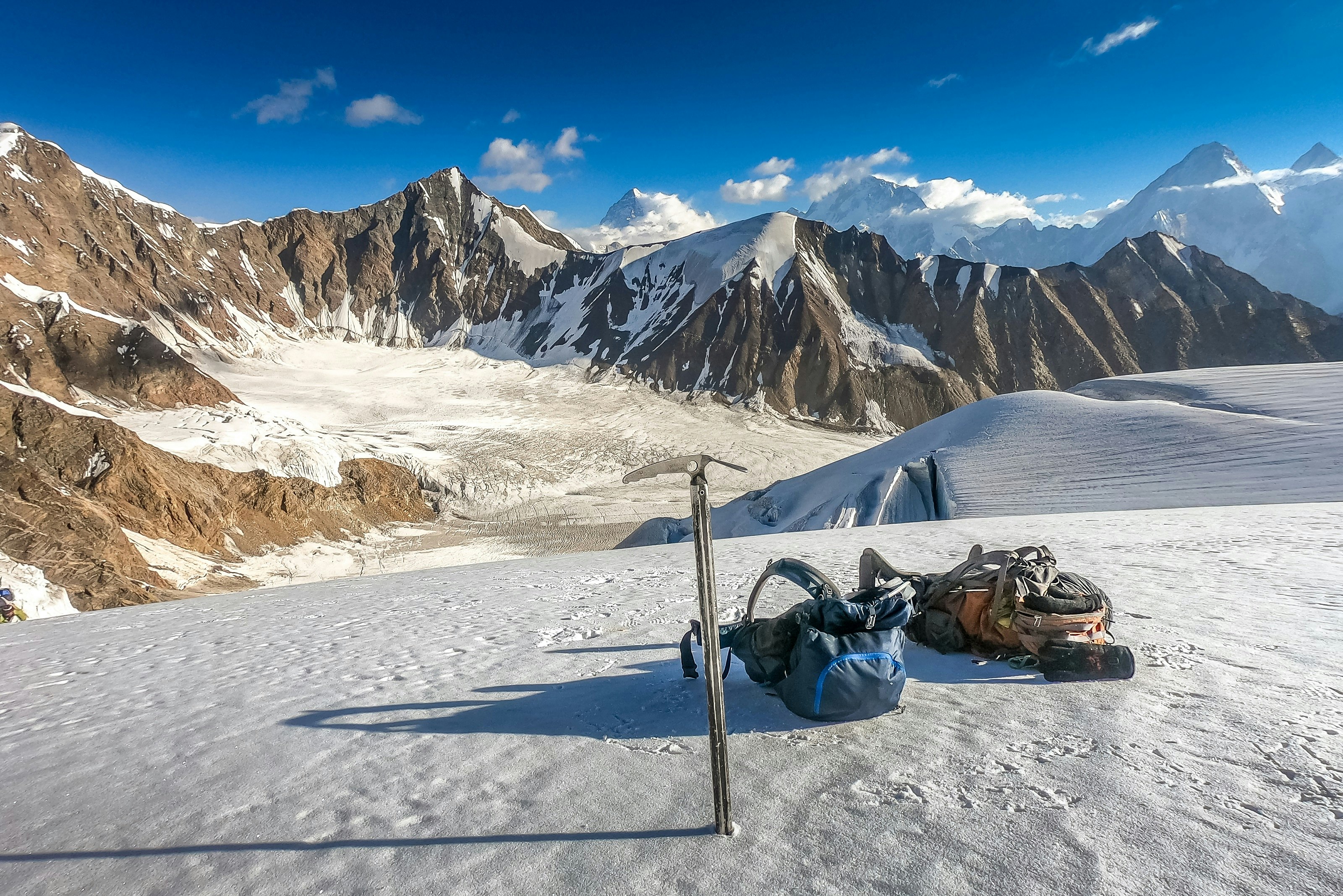 An ice pick is jammed into the ice on the Gondogoro La pass, next to which, two trekking backpacks lie on the ice. In the background, several rocky summits are visible.