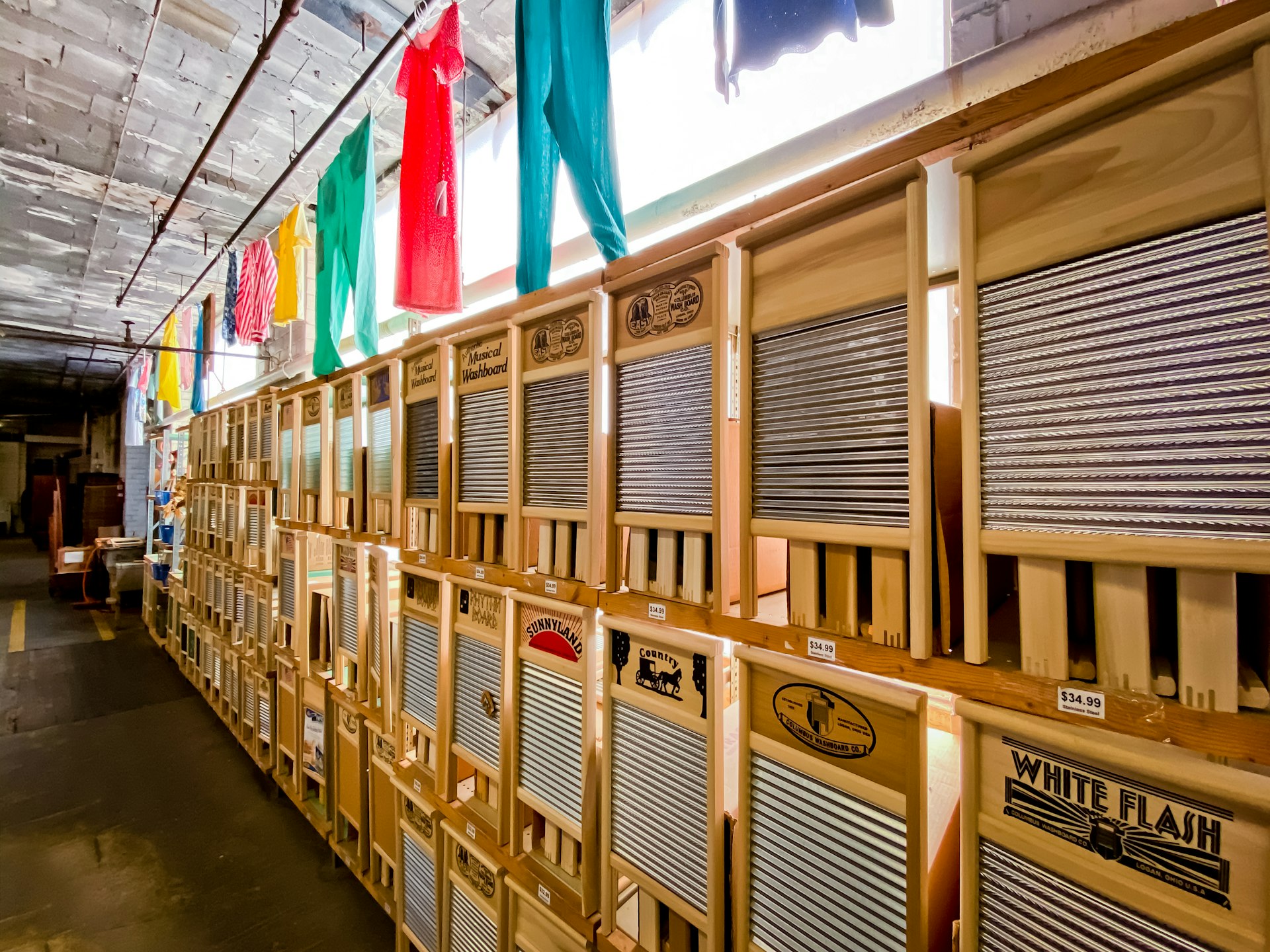 A long row of washboards produced in Logan Ohio is illuminated by a strip of clerestory windows around the top edge of the factory walls. In front of those windows hang brightly colored articles of clothing, like laundry out to dry.