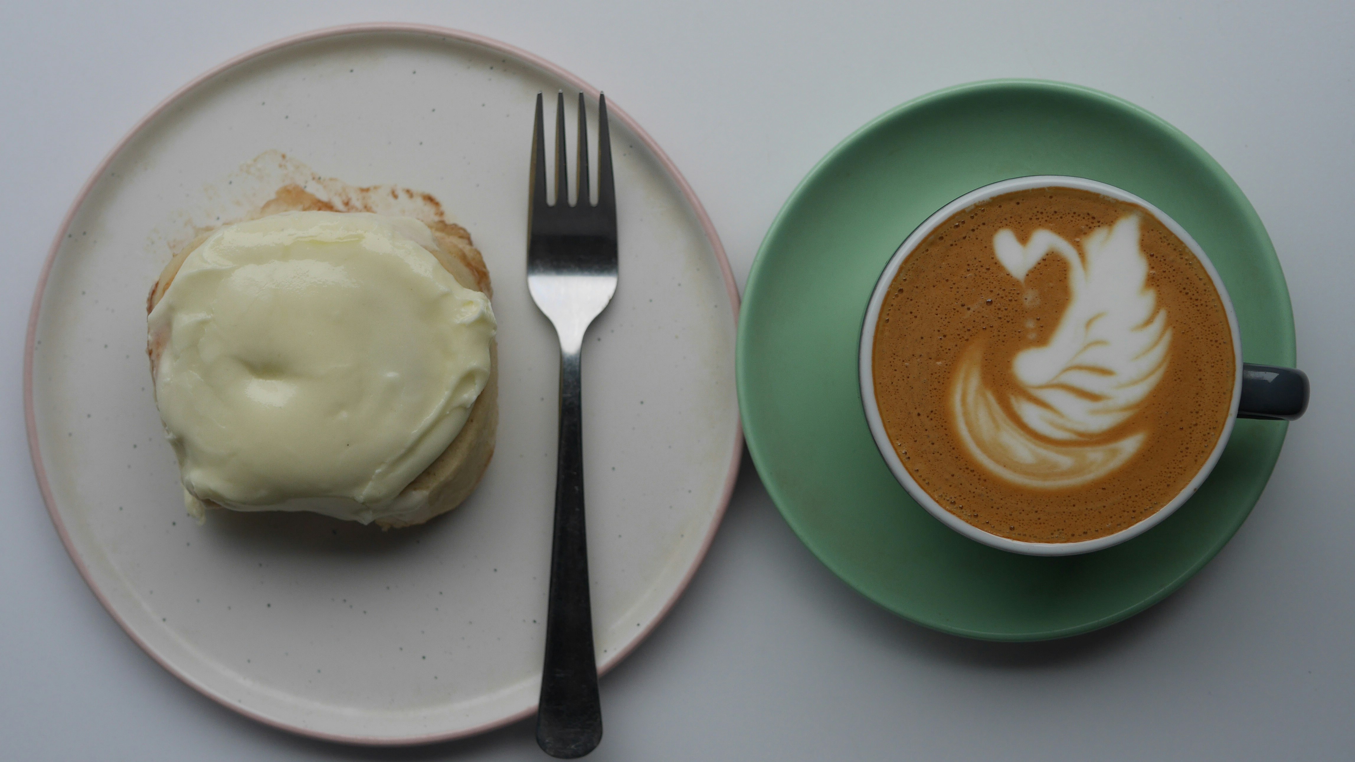 A cake with white icing on a white plate with a fork sits next to a green saucer with a cup of coffee with latte art of a leaf.
