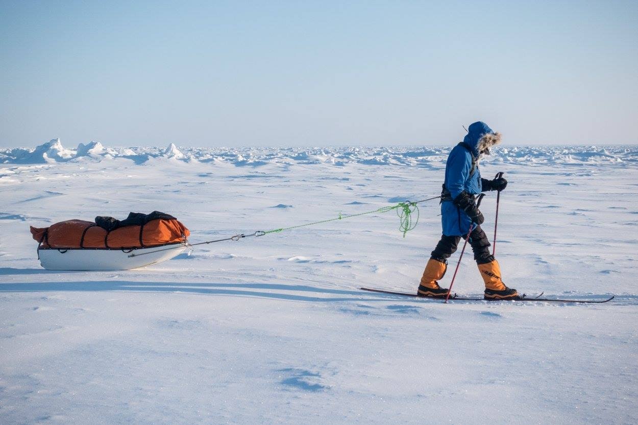 a man in a blue parka walks across ice and snow pulling an orange sled behind him