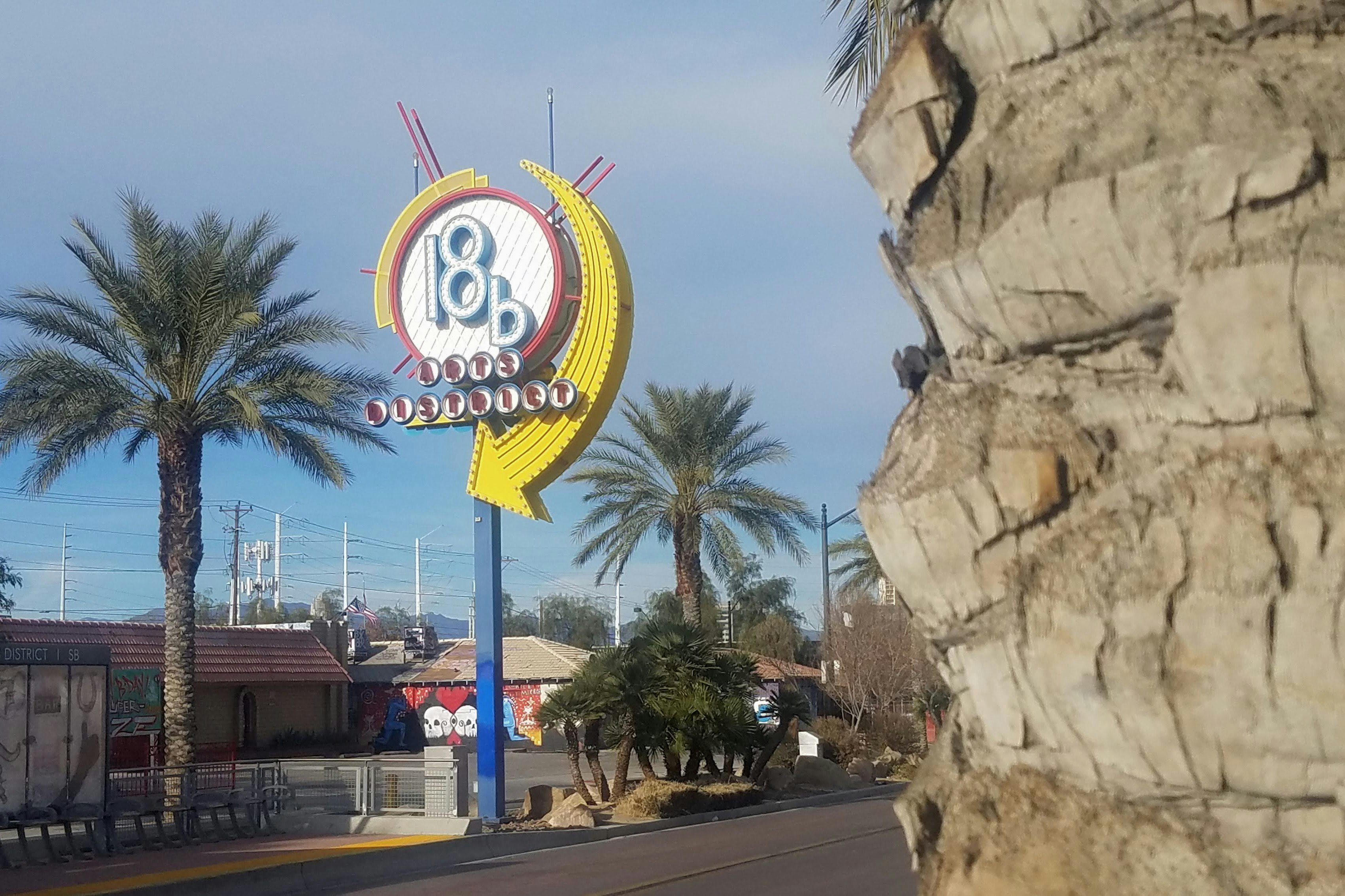 A tall sign, reminiscent of a lollypop, stands beside a palm tree on a street in Las Vegas; the sign states: 18b Arts District.