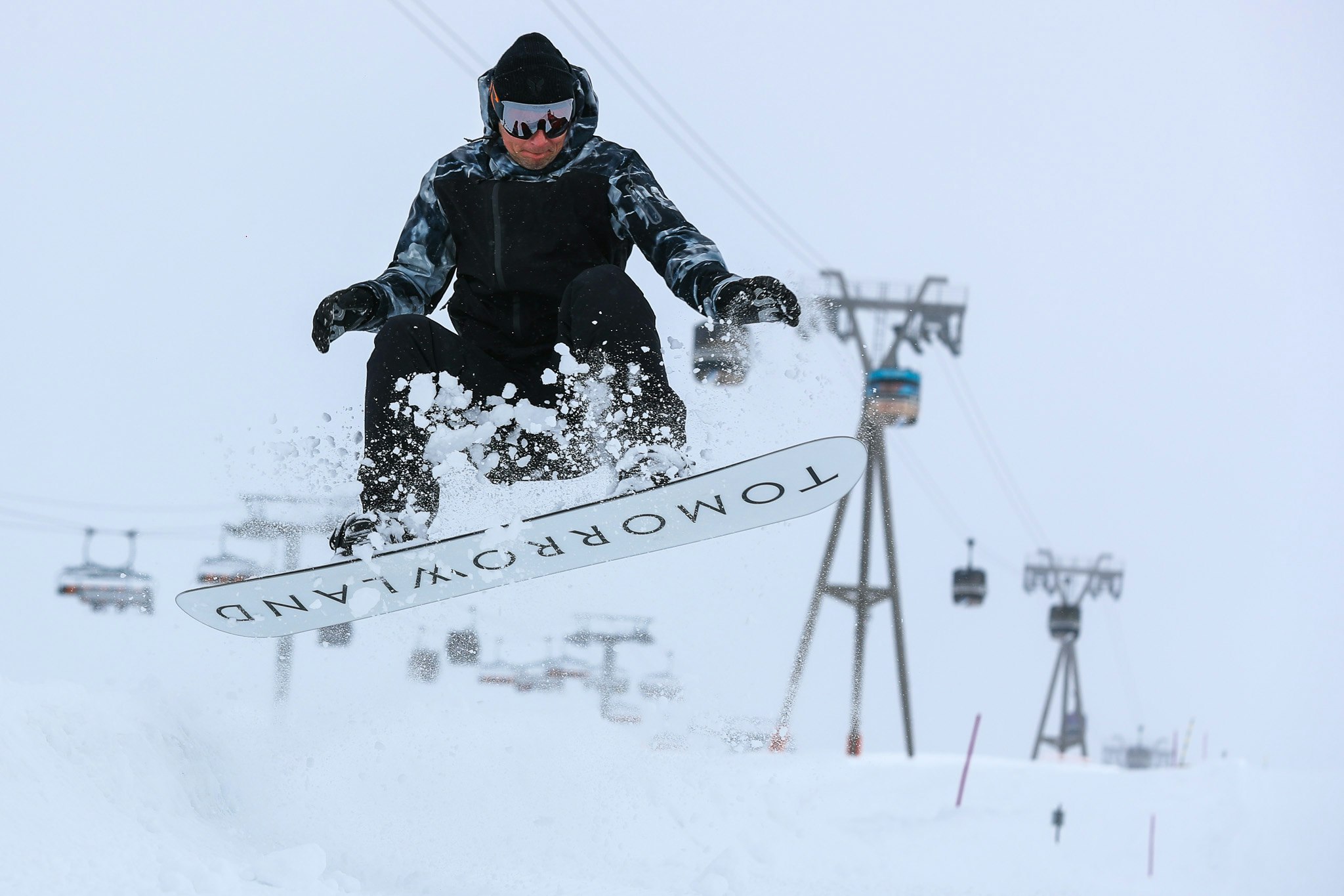 A snowboarder dressed all in black does a jump, exposing the underside of their board which reads 'TOMORROWLAND' at Tomorrowland Winter in Alpe D'Huez, France. 