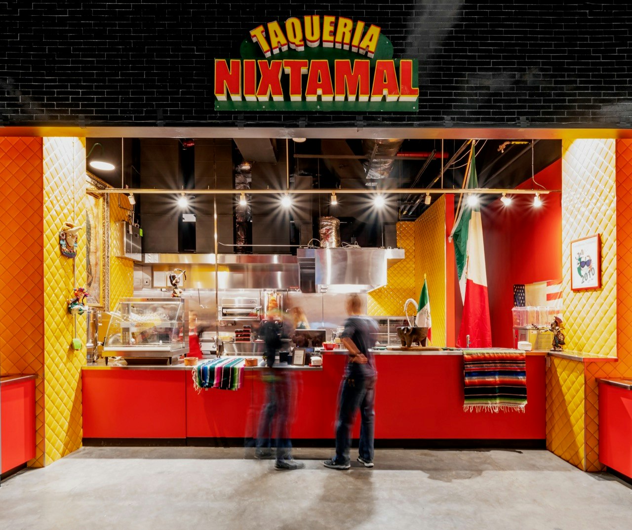 The Taqueria Nixtamal storefront - bright red counter, yellow and red walls, with Mexican flags hanging to the right