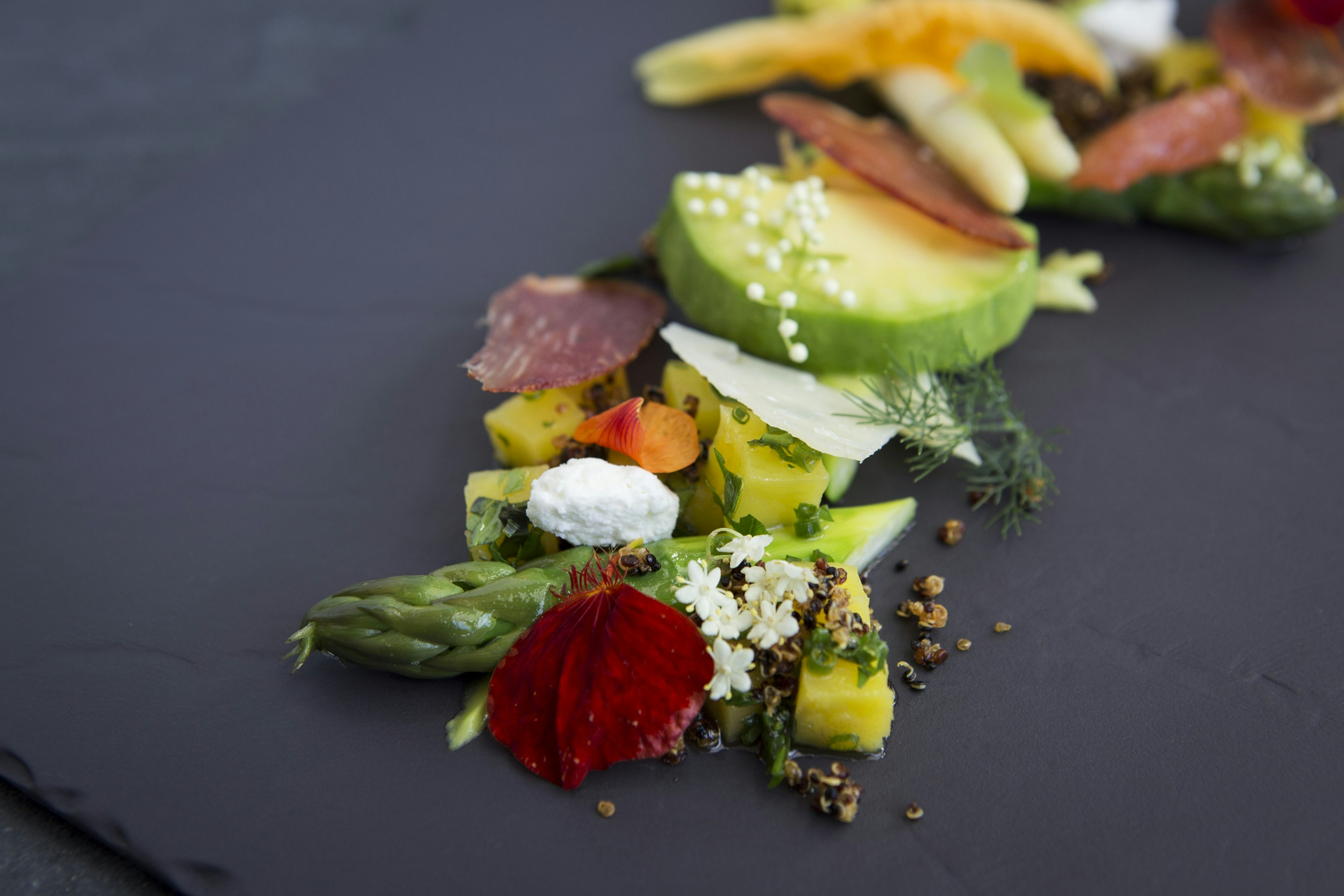 An intricate presentation of colorful vegetables and cured meat on a gray slate.
