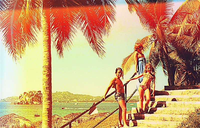 An adult and two young teenagers stand on stone steps beside several large palm trees with the beach and sea behind. The image has an aged feel.