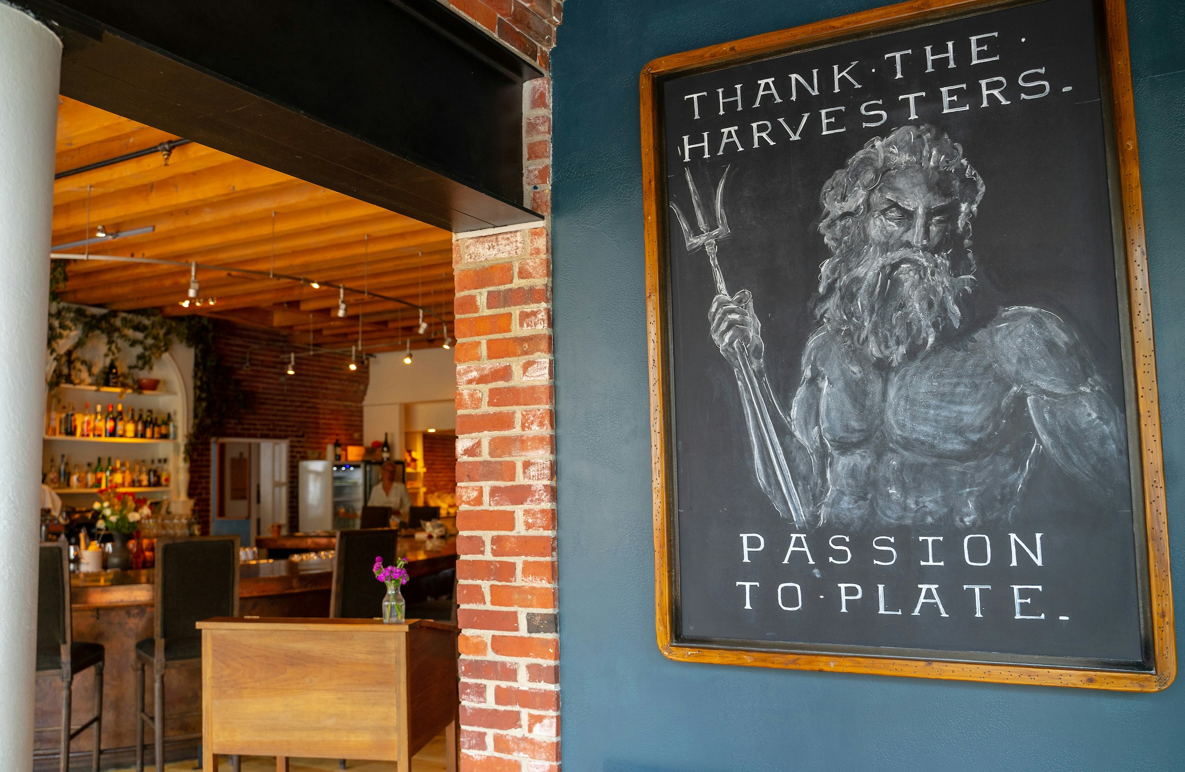 The entrance to Solo Italiano on Portland, Maine features a black chalkboard with a drawing of the sea god Poseidon and the words "Thank the Harvesters" "Passion to Plate" in a serif font. Beyond is a warm room in brick and wood with a brightly lit bar full of colorful bottles and a hostess stand with pink flowers in a bud vase 
