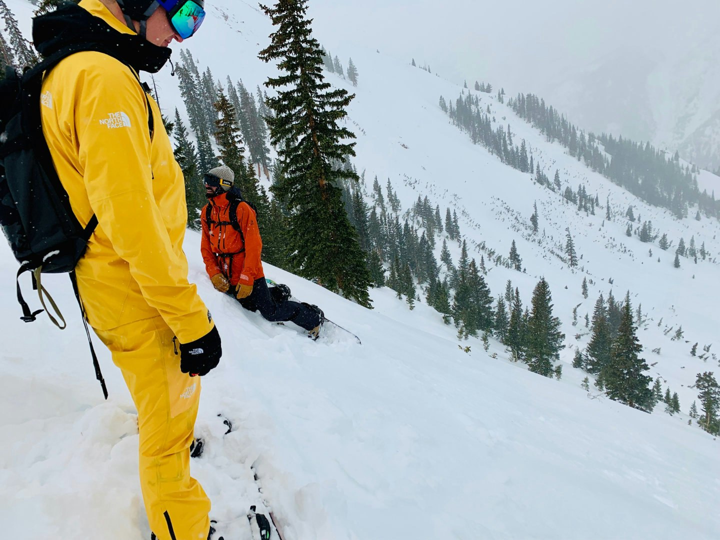 Two skiers peer down a steep slope in Aspen, Colorado with evergreen trees in the background. The skiiers wear bright yellow FutureLight pants and jackets by North Face, along with black backpacks, gloves, and goggles.