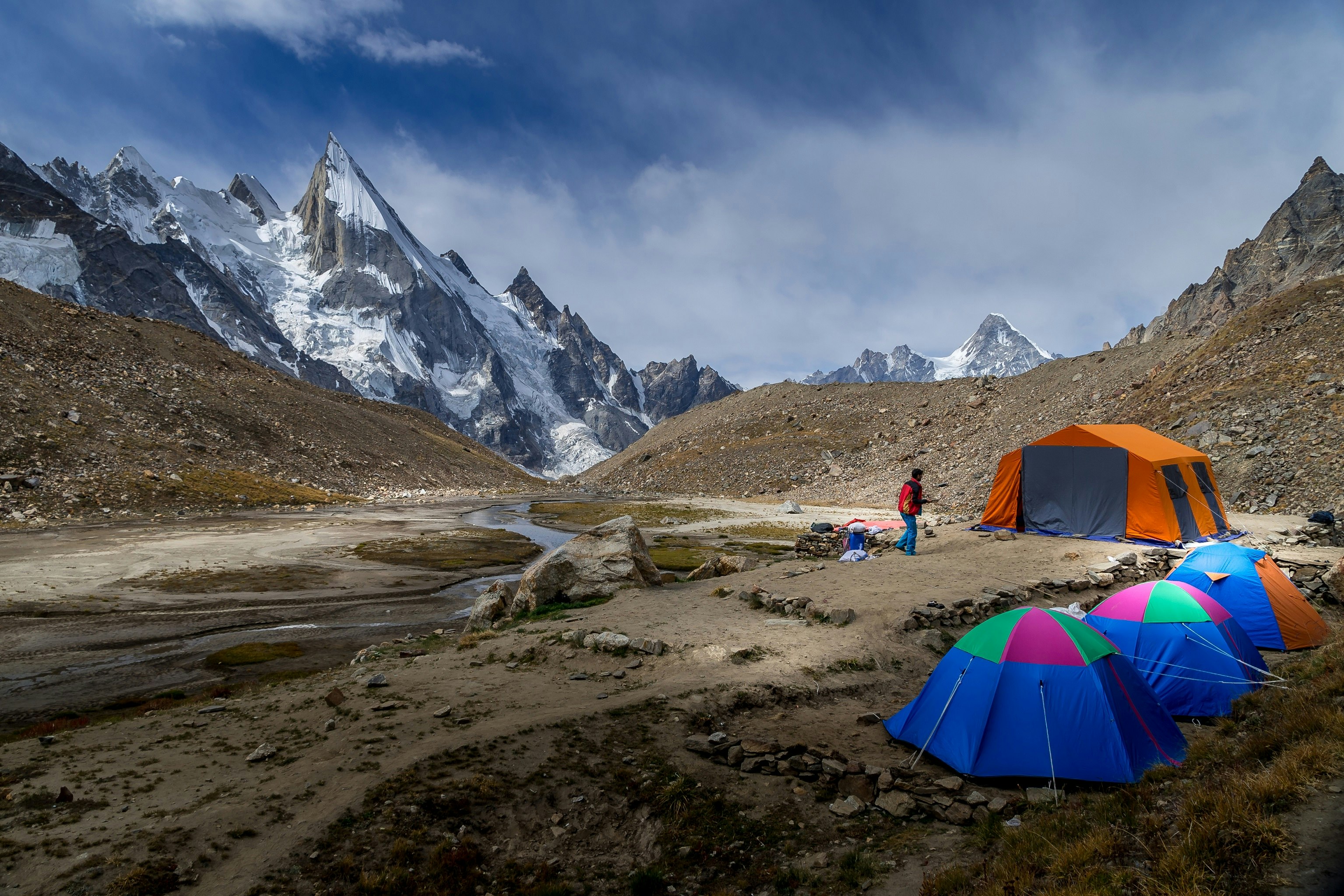 A series of colourful tents stand in a line at the campsite in Lalia Peak, part of the K2 base camp trek. A narrow river runs past the camp site, while a number of jagged peaks are visible in the background.