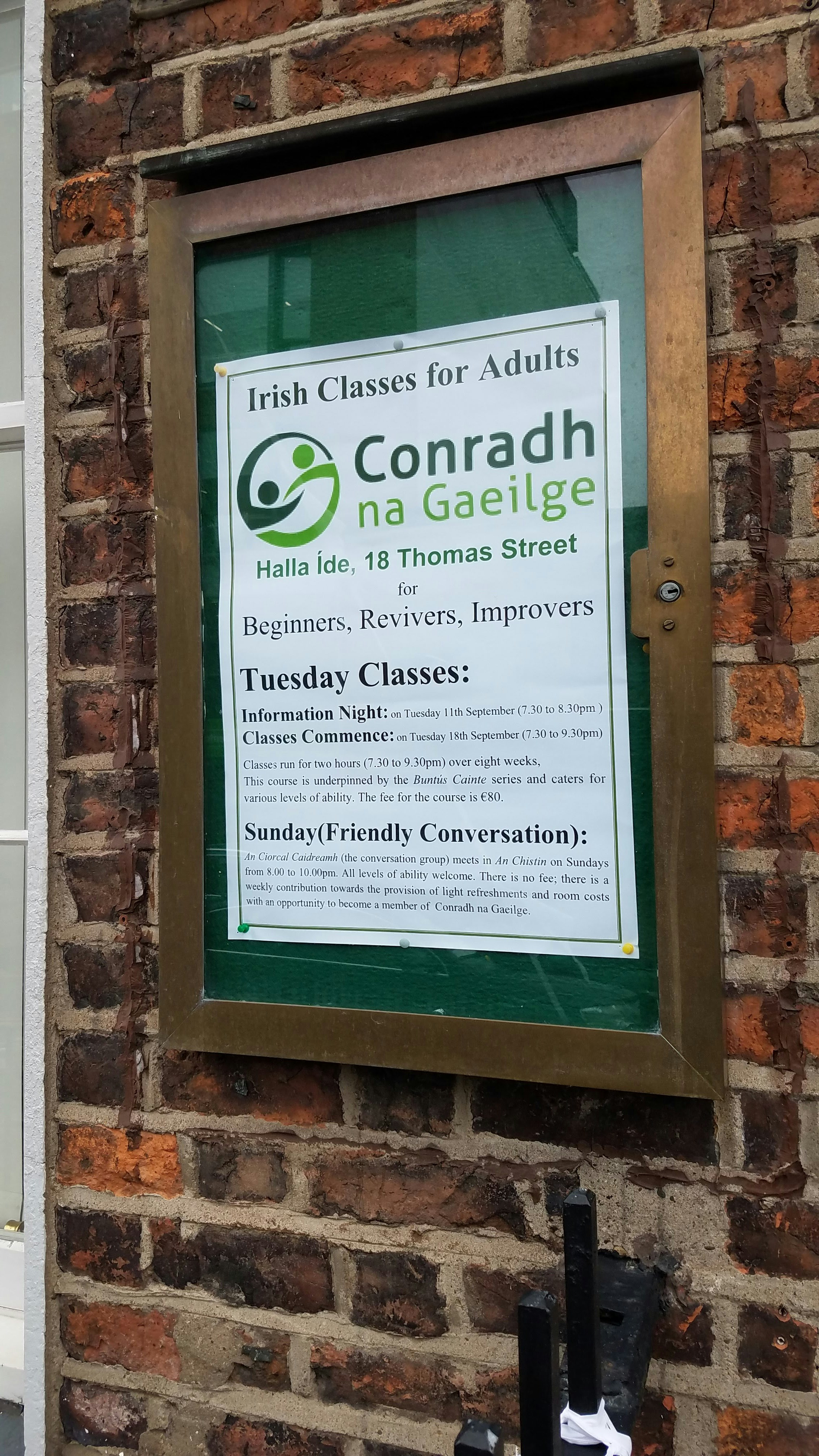 A sign advertising Conradh na Gaeilge, or Irish Classes for Adults" in Limerick hangs in a metal and glass poster box against a green background on a brick wall
