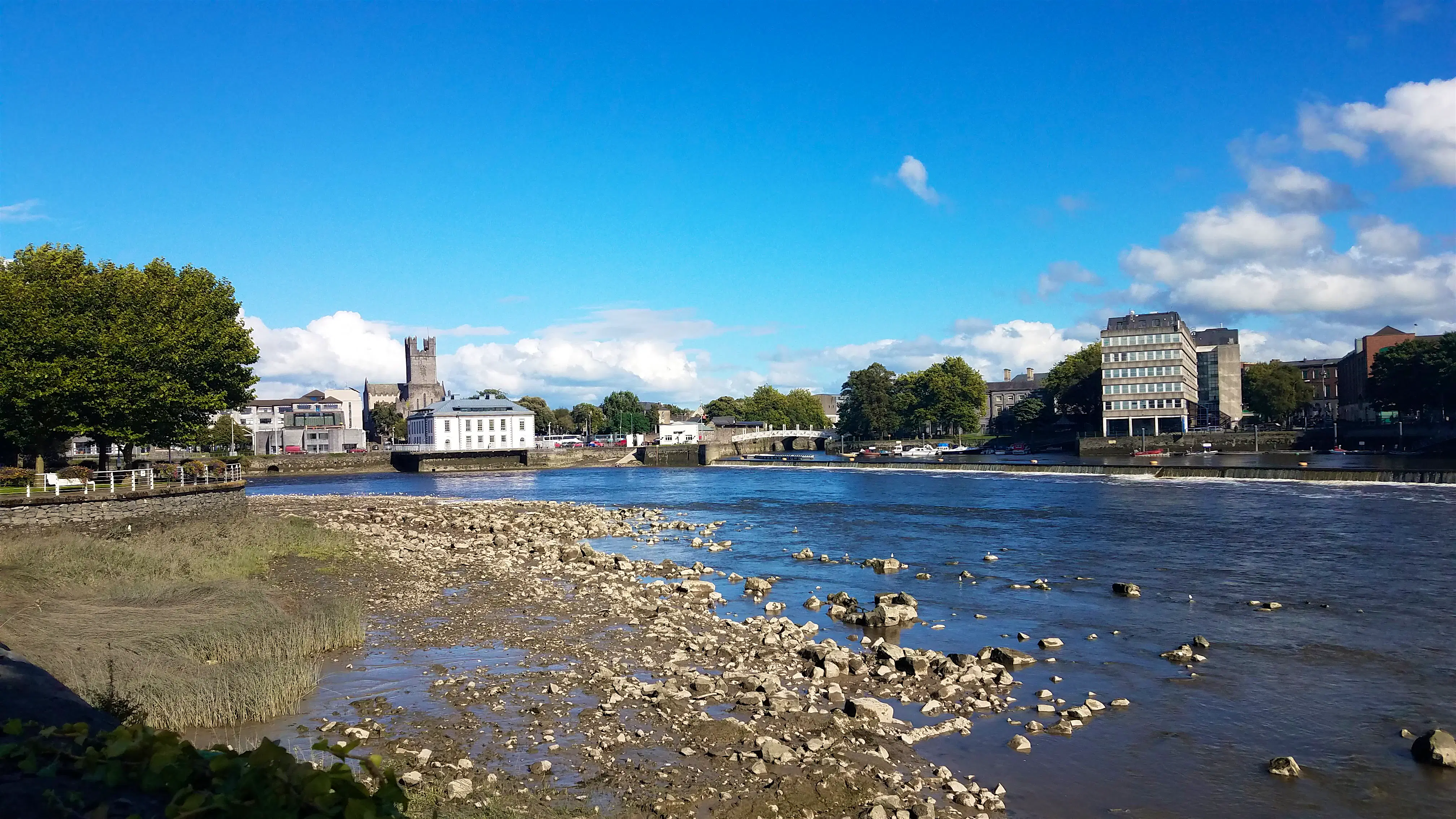 river-shannon-limerick.jpg The river Shannon's rocky shore gives way to deep blue water reflecting the lighter, brighter blue of the sky overhead. On the opposite shore are stone buildings full of glass windows, a bridge, and in the distance, a crenelated medieval tower.
