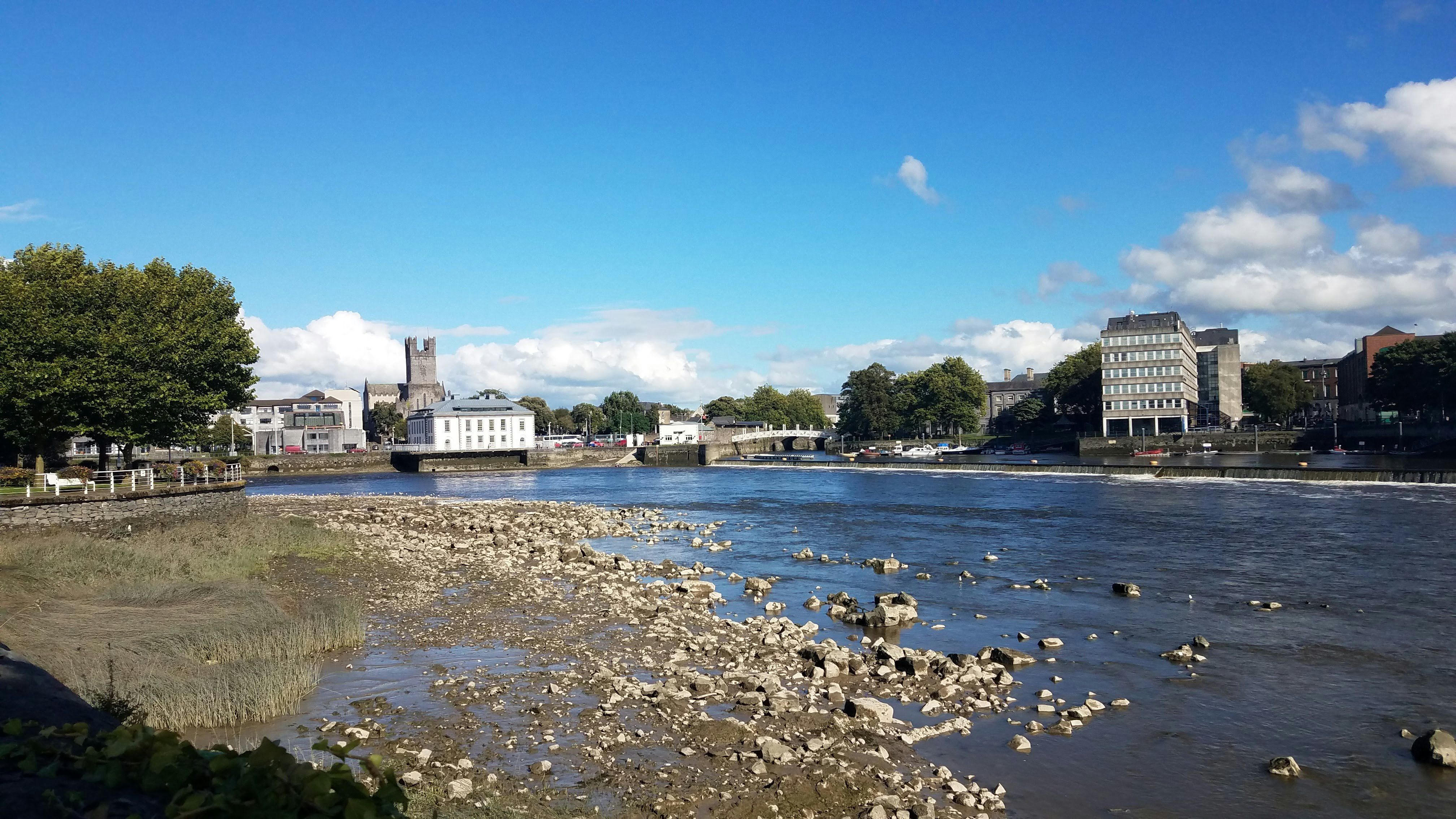 The river Shannon's rocky shore gives way to deep blue water reflecting the lighter, brighter blue of the sky overhead. On the opposite shore are stone buildings full of glass windows, a bridge, and in the distance, a crenelated medieval tower. 
